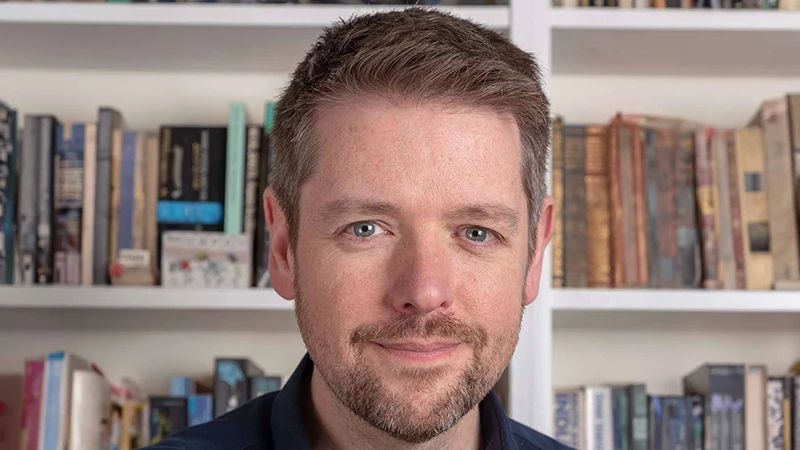 Portrait of children’s author Alastair Chisholm in front of bookshelves