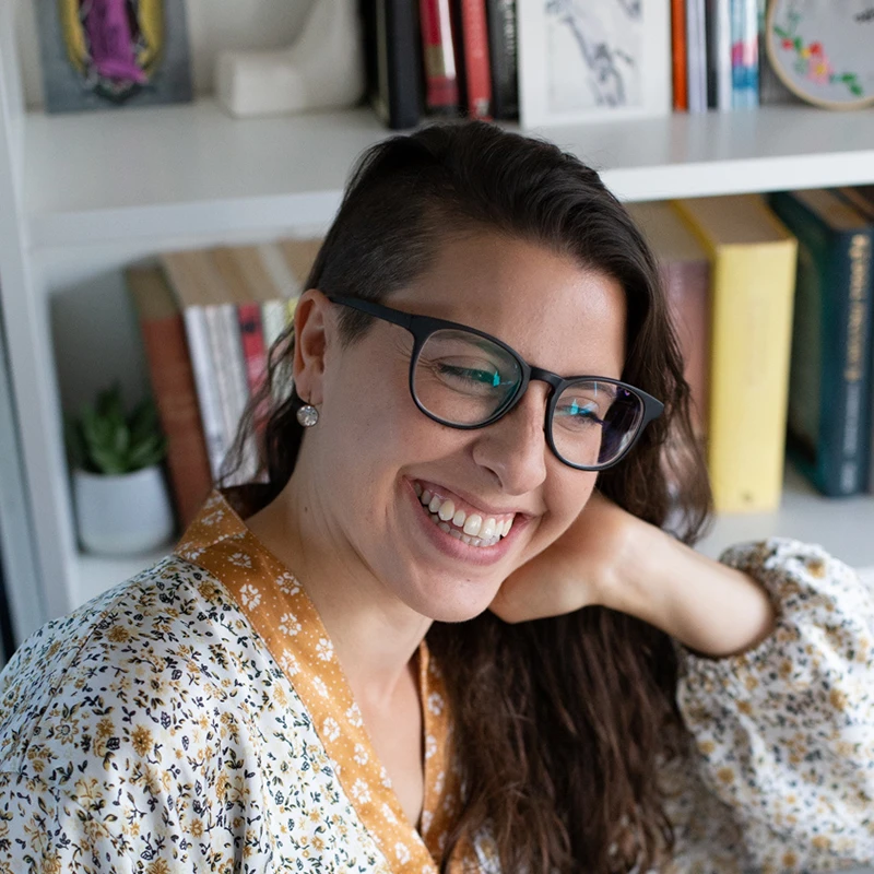 Portrait of Angie Spoto smiling while holding an open book in front of a bookshelf.