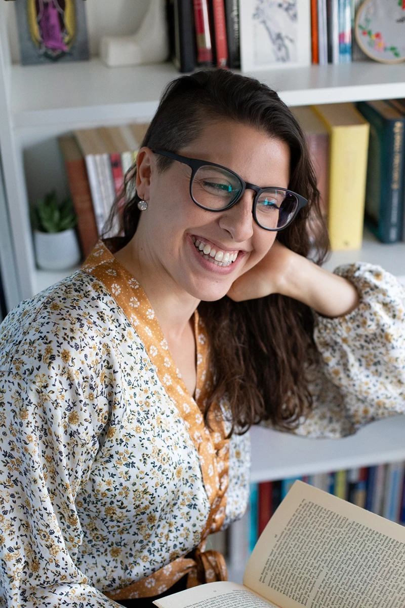 Portrait of Angie Spoto smiling while holding an open book in front of a bookshelf.