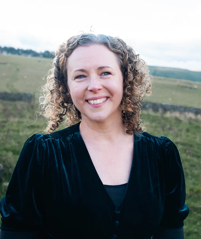 Anna Caig standing outdoors in a rural landscape, smiling, with rolling hills in the background.