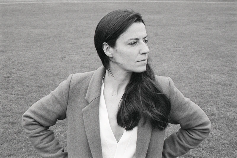 Black-and-white portrait of Cal Flyn standing outdoors on grass, looking to the side with hands on hips and wearing a blazer.