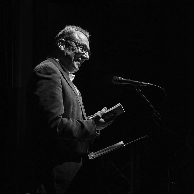Jim Carruth reading from a book at a microphone on stage in a black-and-white photo.