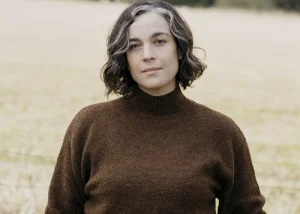 Kristie De Garis standing outdoors in a field, wearing a brown jumper, with trees in the background.