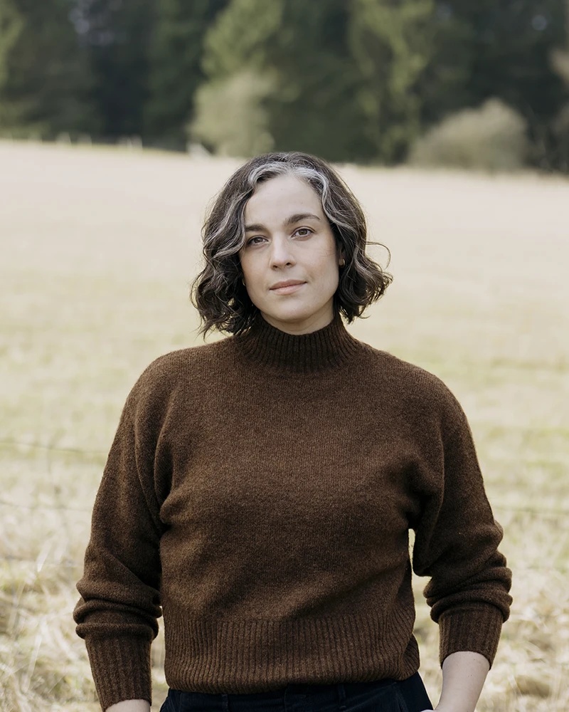 Kristie De Garis standing outdoors in a field, wearing a brown jumper, with trees in the background.