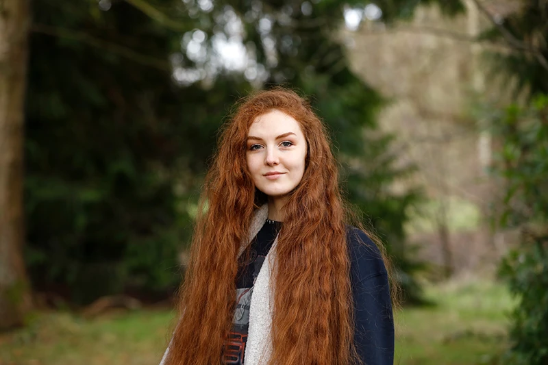 Portrait of Len Pennie standing outdoors in a leafy setting, with long red hair and wearing a dark top and scarf.