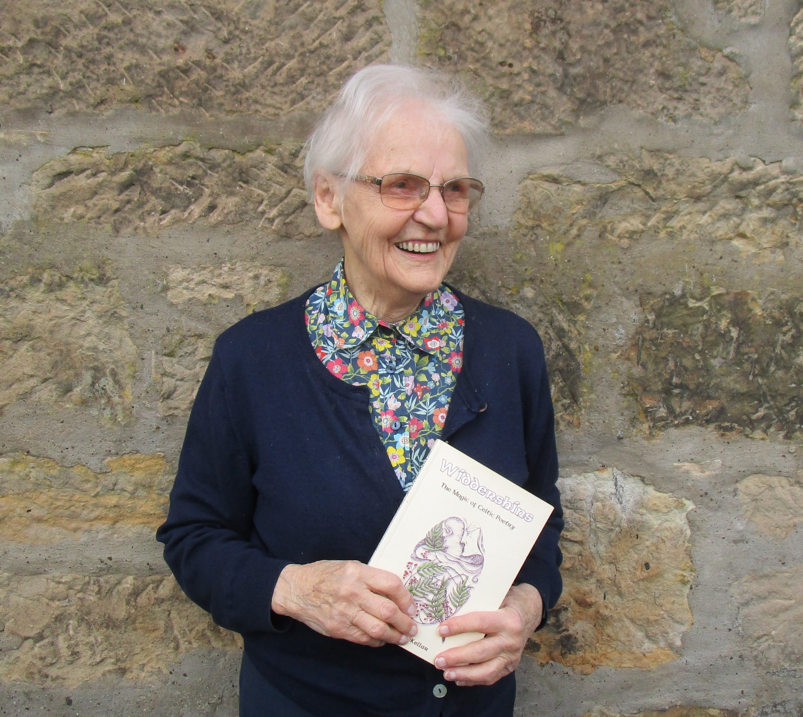 Betty McKellar standing outdoors against a stone wall, smiling and holding a copy of her book Widdershins.