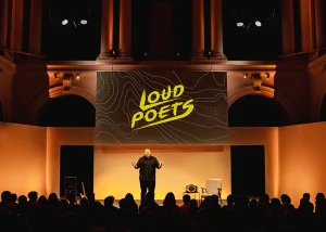 Performer on stage beneath a large “Loud Poets” screen at Paisley Town Hall, with an audience seated in front of the stage.