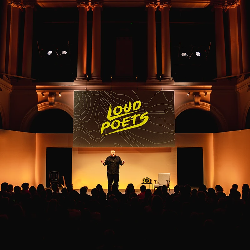Performer on stage beneath a large “Loud Poets” screen at Paisley Town Hall, with an audience seated in front of the stage.