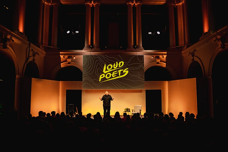 Performer on stage beneath a large “Loud Poets” screen at Paisley Town Hall, with an audience seated in front of the stage.