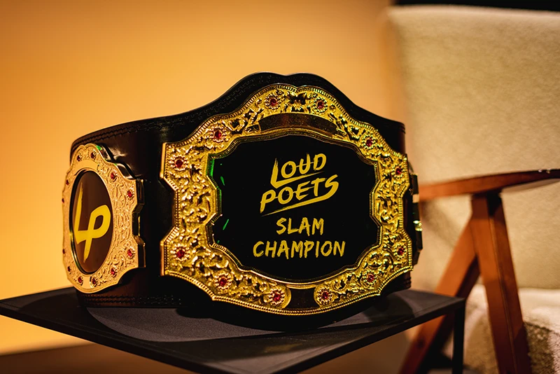 Championship belt engraved “Loud Poets Slam Champion” resting on a chair under warm stage lighting.