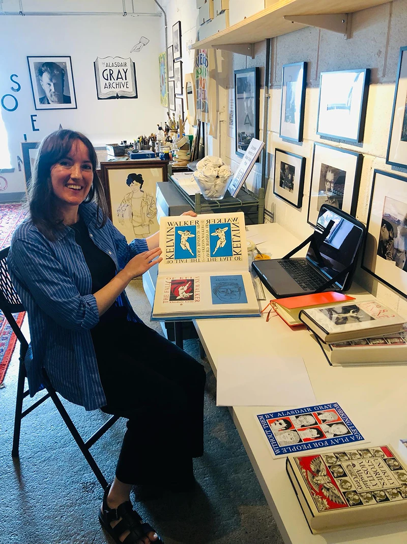 Black-and-white portrait of Maeve Redmond standing beside signage for the Alasdair Gray Archive.