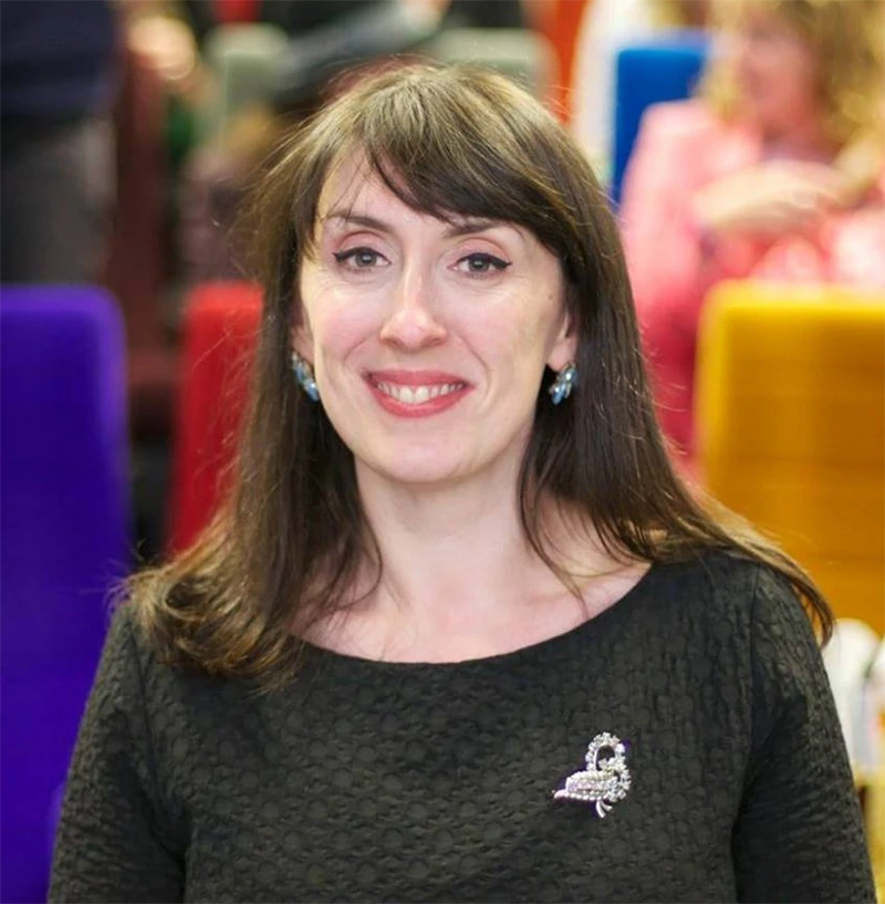 Portrait of Mairi Kidd smiling indoors against a colourful blurred background.