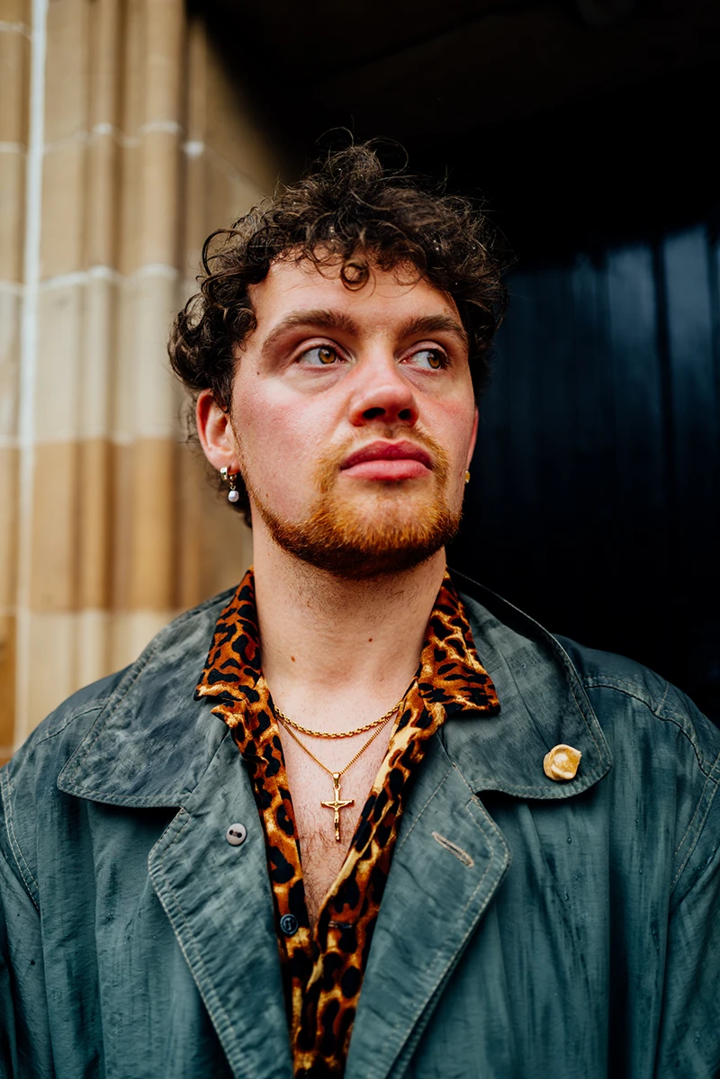 Portrait of Michael Mullen standing outdoors beside a stone building, wearing a green coat and patterned shirt, looking off to the side.