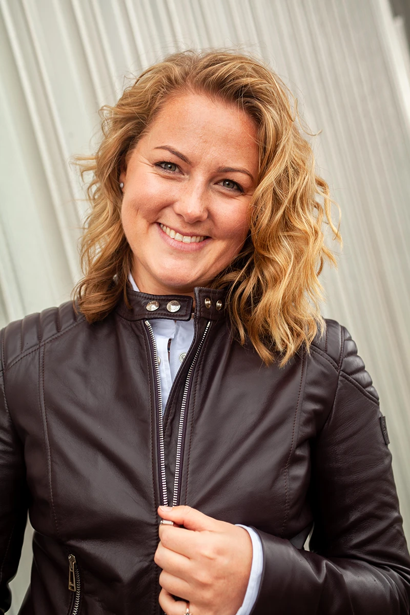 Portrait of Mollie Hughes smiling at the camera, wearing a brown leather jacket and standing in front of a light-coloured wall.