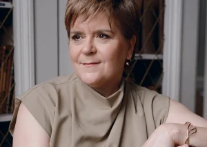 Portrait of Nicola Sturgeon seated indoors, leaning on the back of a chair and looking to the side, wearing a sleeveless beige top.