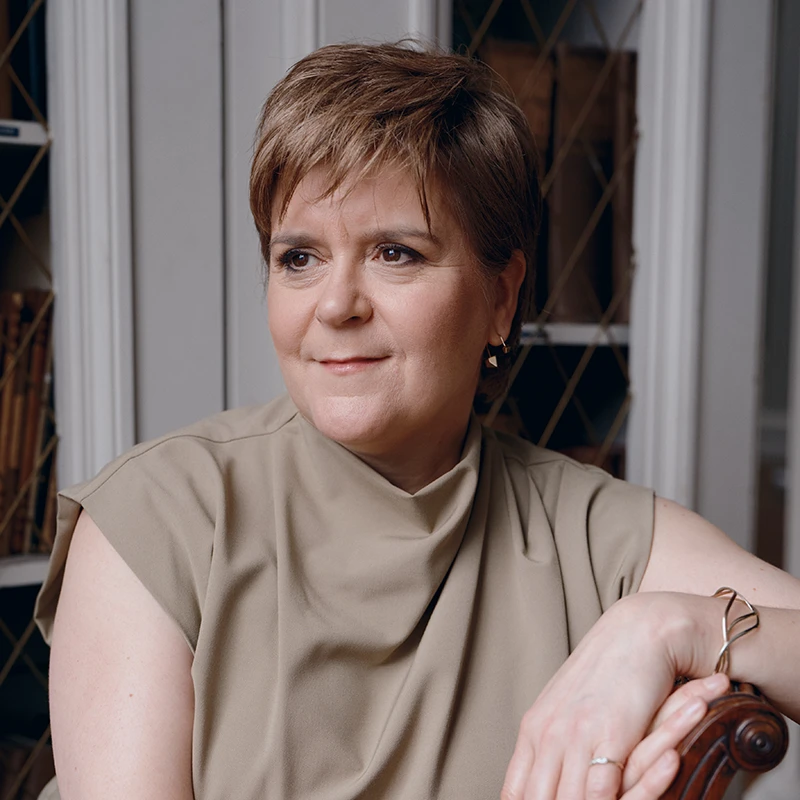 Portrait of Nicola Sturgeon seated indoors, leaning on the back of a chair and looking to the side, wearing a sleeveless beige top.