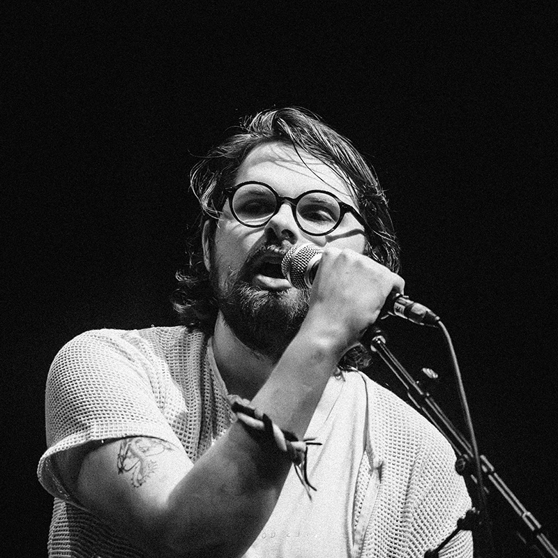 Black and white photo of Robbie MacLeòid performing on stage, singing into a microphone.