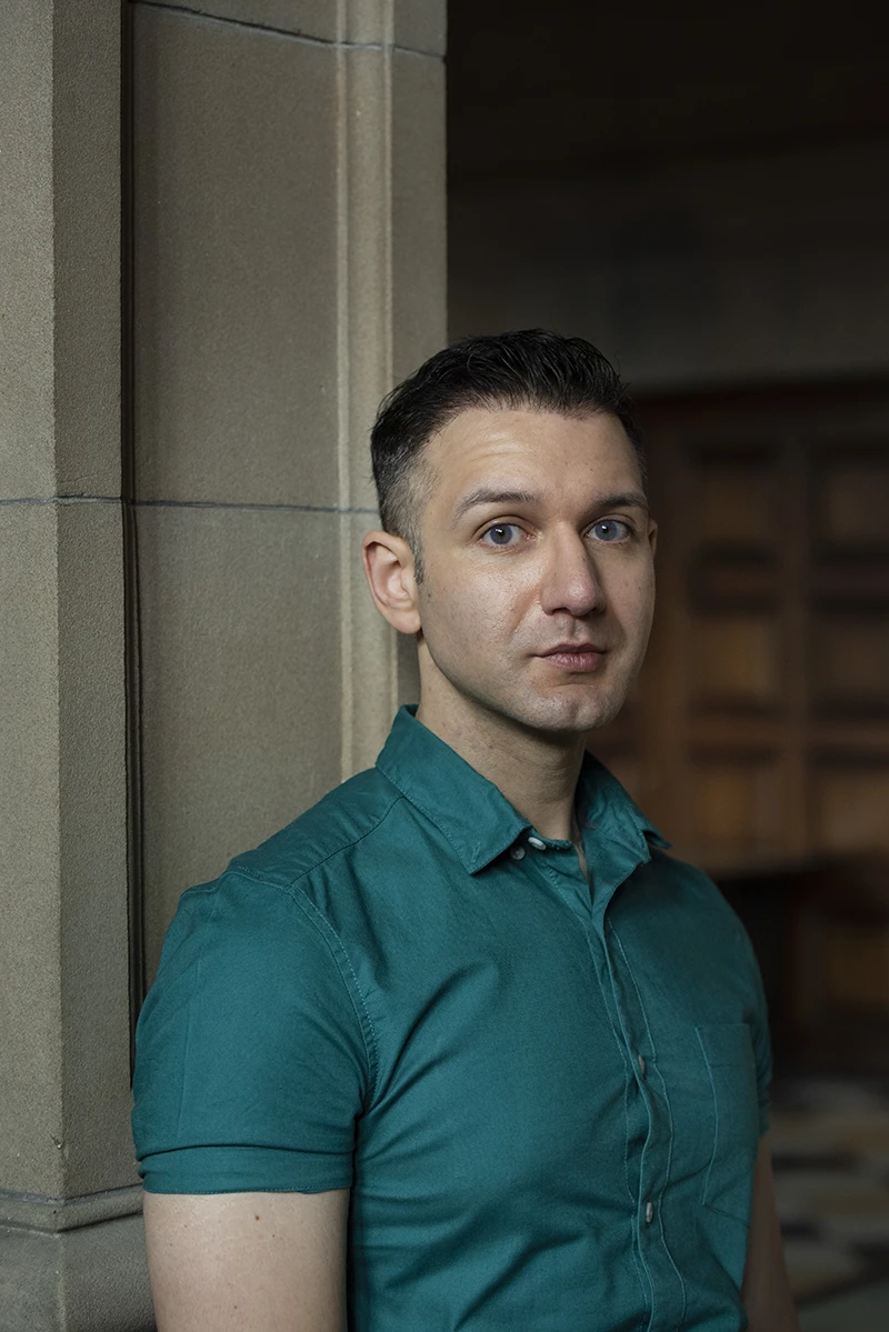 Steven Veerapen standing indoors beside a stone pillar, wearing a green shirt and looking towards the camera.