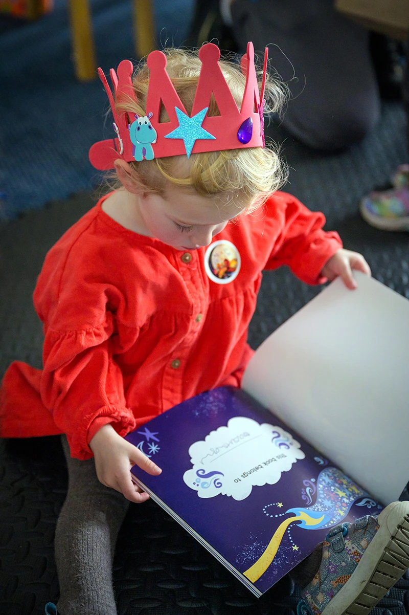 Young child wearing a paper crown reading a picture book during a storytelling event.