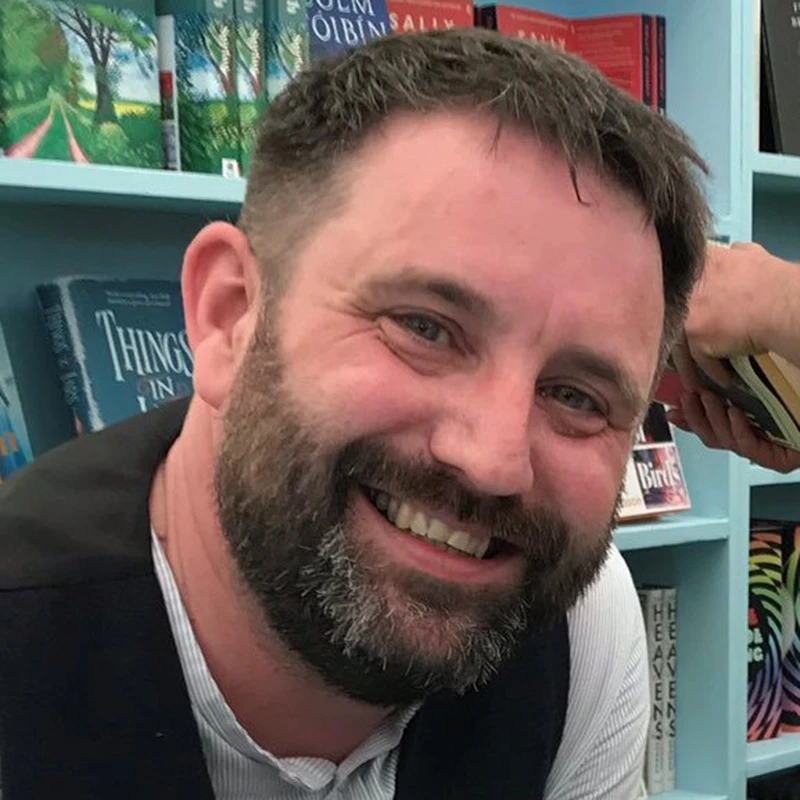 Chae Strathie smiling in a bookshop with shelves of books behind him.