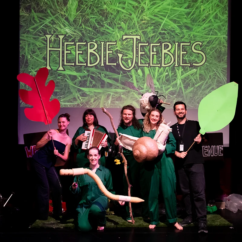 Performers posing on stage with oversized leaf props and insect puppets from the children's show Heebie Jeebies.