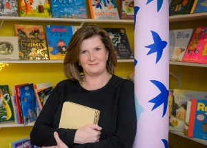 Author Rachel Huggins holding a book while standing in front of shelves of children's books.