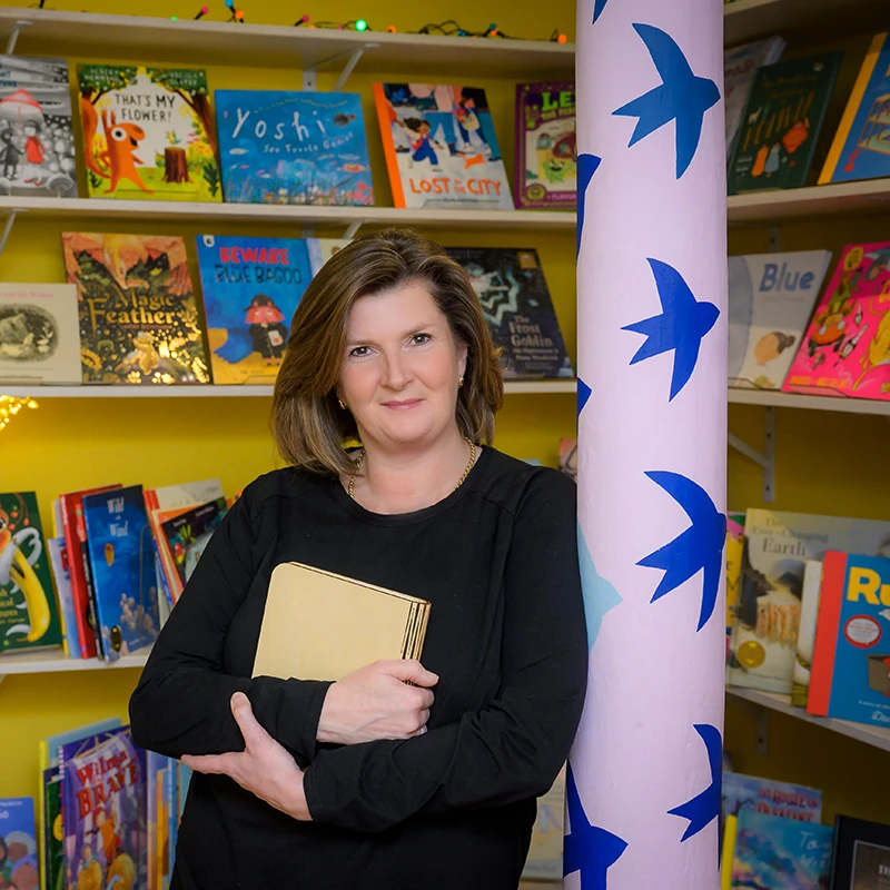 Author Rachel Huggins holding a book while standing in front of shelves of children's books.