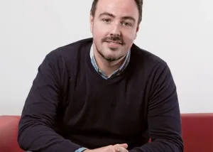 Ross MacKenzie smiling while seated on a red sofa.