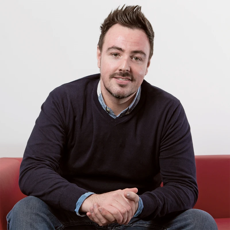 Ross MacKenzie smiling while seated on a red sofa.