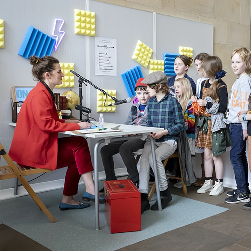 Performer interviewing a young child at a microphone in the colourful Experts Radio Lab studio.