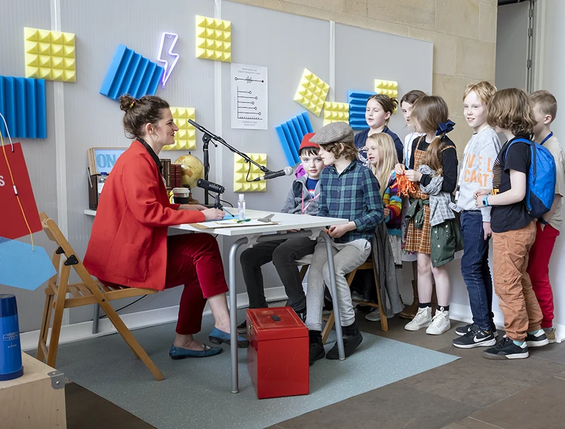 Performer interviewing a young child at a microphone in the colourful Experts Radio Lab studio.